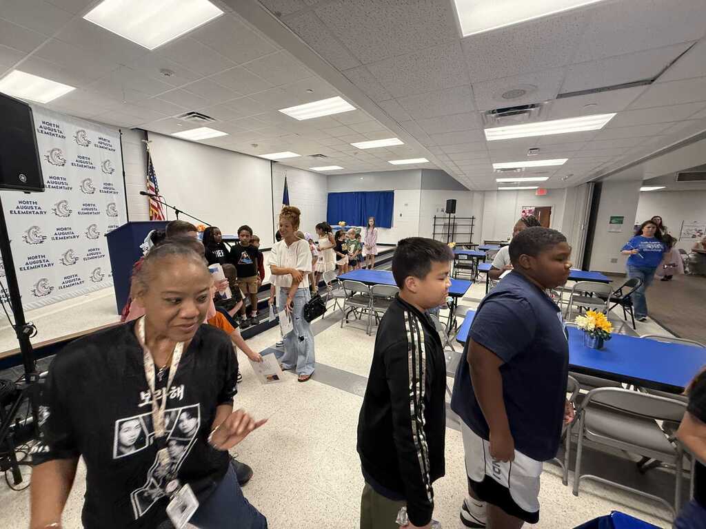 A classroom with tables and chairs. Two adults and three children stand around. One adult wears a lanyard.
