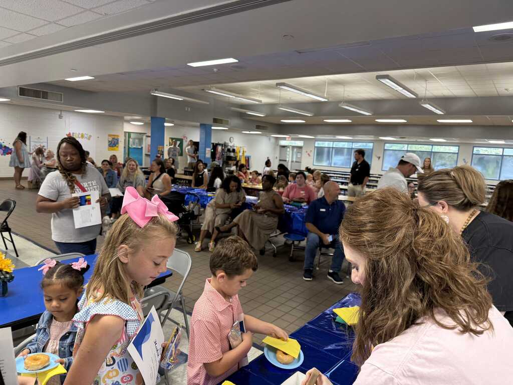 A group of children and adults are sitting at tables, possibly at an event. Tables have blue tablecloths and plates of food.