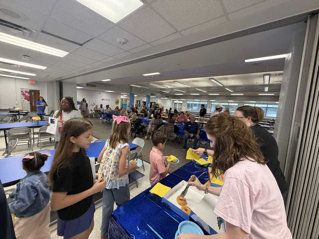 Indoor dining area with children and adults. A group stands around a blue table, with a person cutting food.
