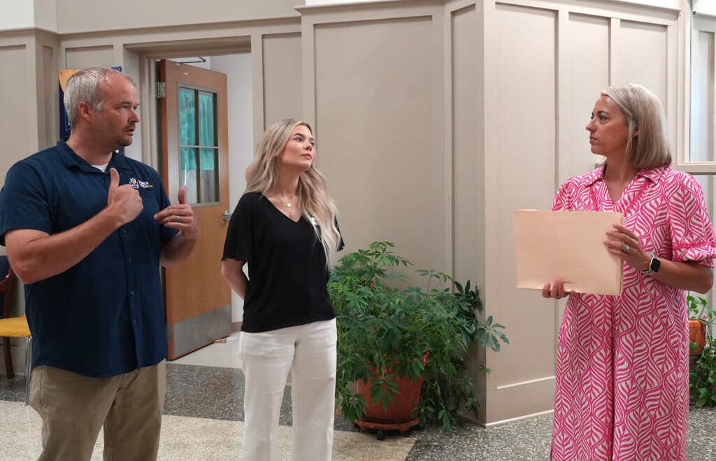 Three adults stand in a room. A man in a blue shirt gestures with both hands. A woman holds a piece of paper.