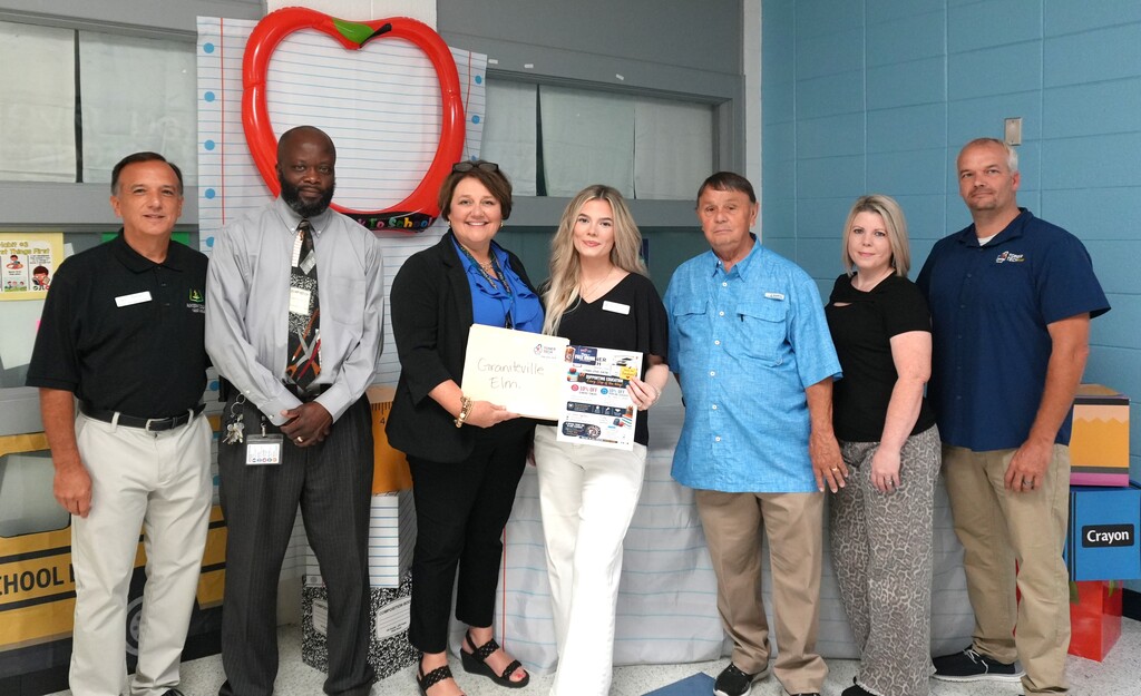 Six people stand near a red apple decoration. A woman holds a certificate, while the others look on.