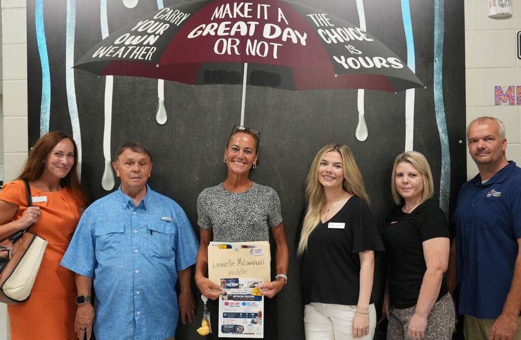 Five adults stand in front of a chalkboard with a mural, holding a certificate and smiling.