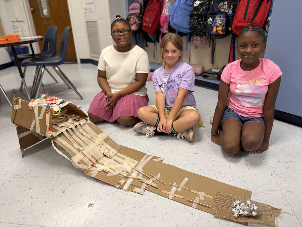 Three girls sit on a floor with a cardboard ramp in front of them. One wears a white shirt and pink skirt. Another wears a pink shirt. A third wears a purple shirt.