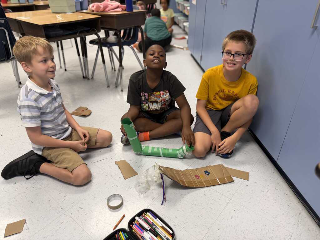 Three children sitting on a floor, engaged in a school activity with a cardboard tube.