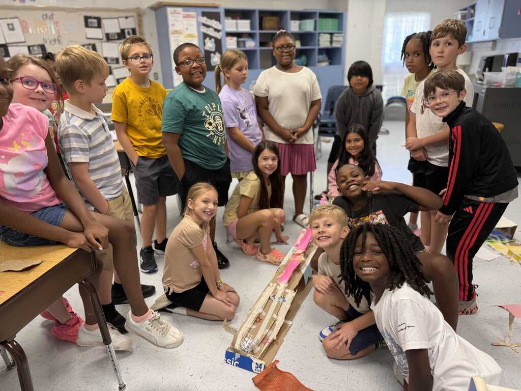 Group of students in a classroom setting, some sitting, some standing, gathered around a cardboard model.