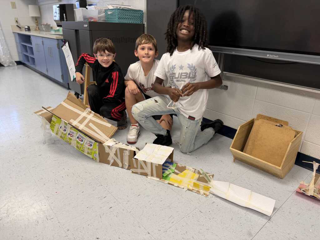Three children pose next to a cardboard project in a classroom. The two on the left hold cardboard, one kneeling on the floor.