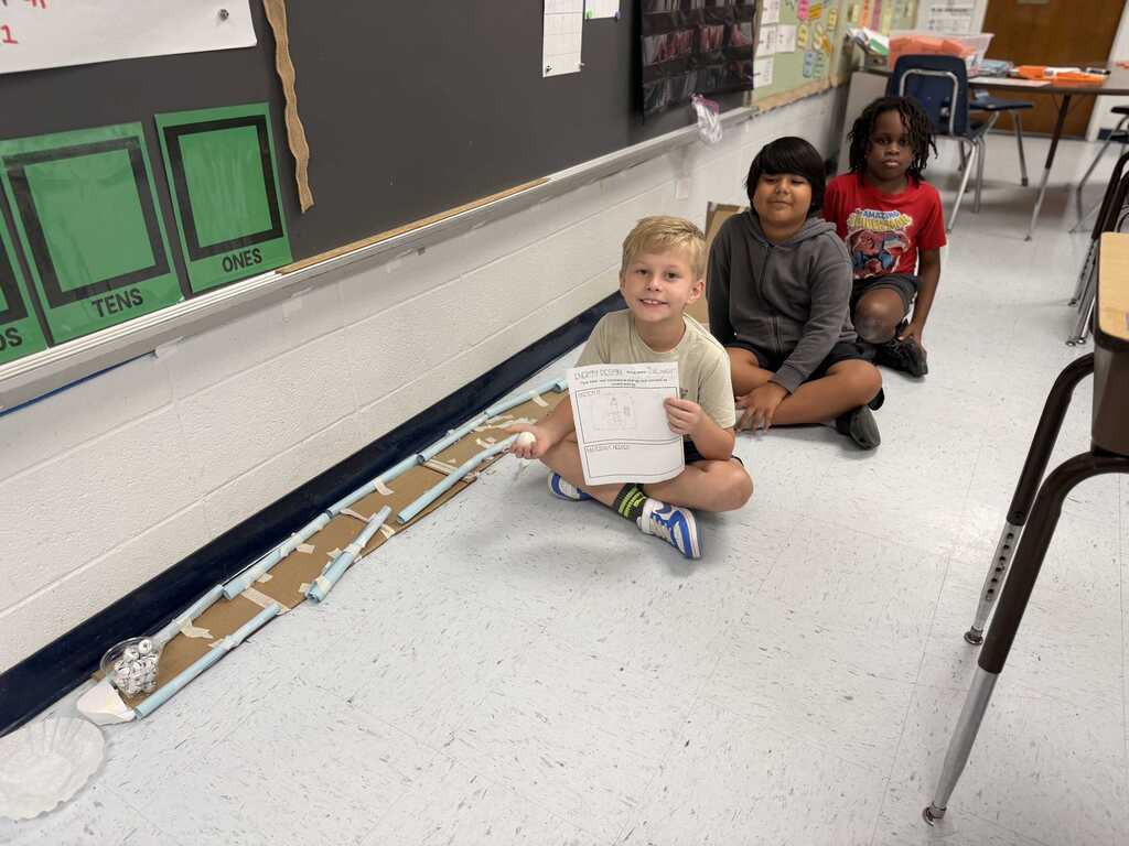 Three children sit on a floor with a long cardboard track. One holds a paper. Behind, a classroom has a blackboard with green boxes.