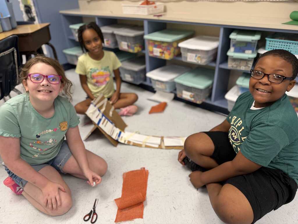 Three children are sitting on the floor in a classroom. Two are smiling, one with glasses, scissors, and orange paper. Behind them are shelves with storage boxes.