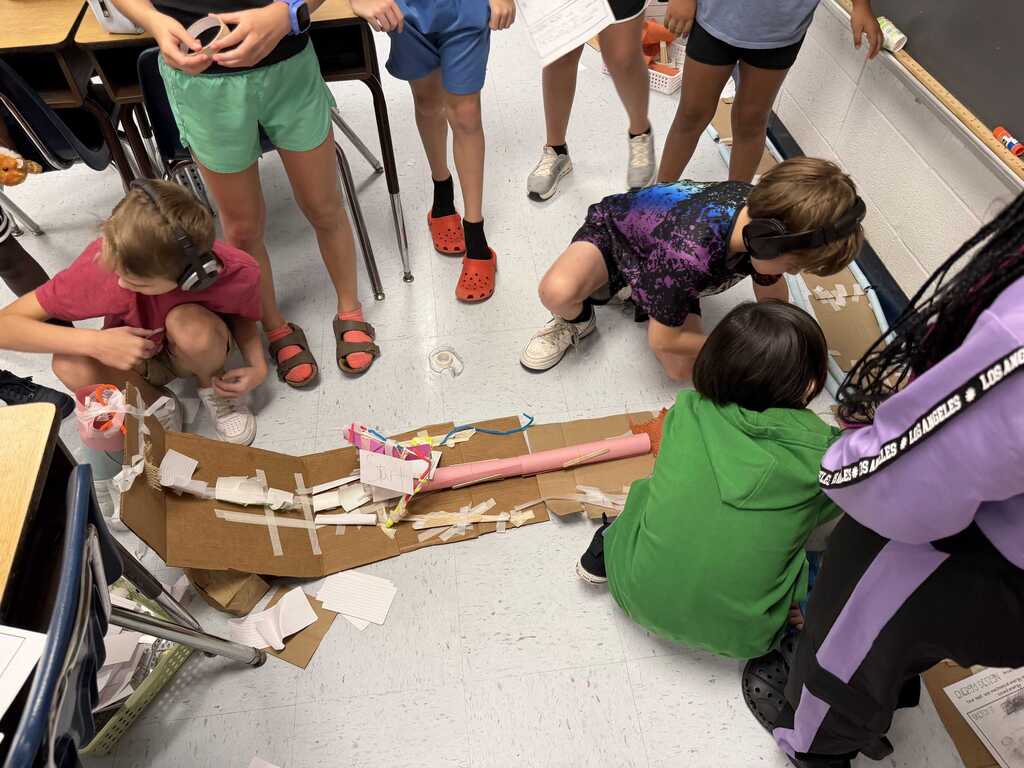 Children gather around a cardboard structure in a classroom, with one student holding a piece of cardboard.