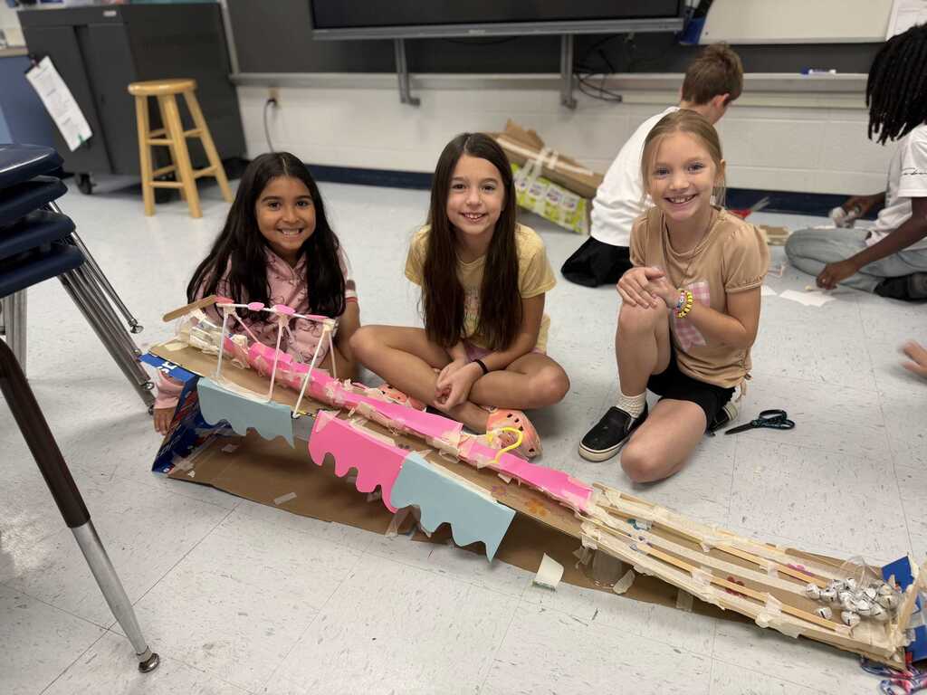 Three girls in a classroom with a cardboard bridge in front of them, smiling. Other students work behind them.