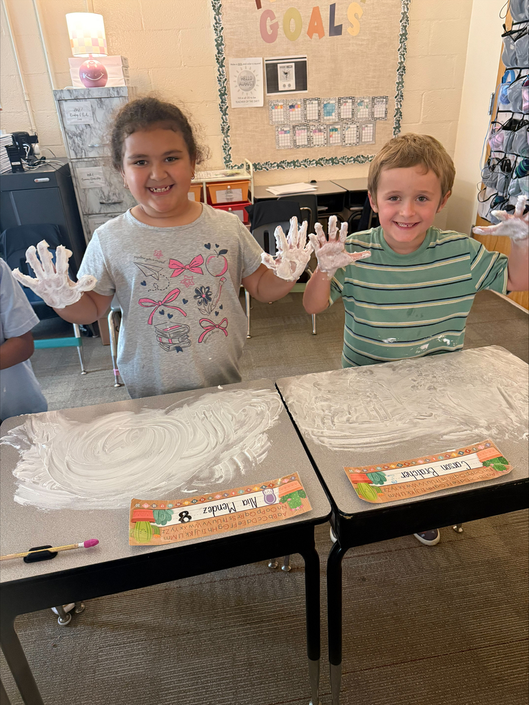 A boy and girl standing behind their desks while holding up their hands that are covered in shaving cream.