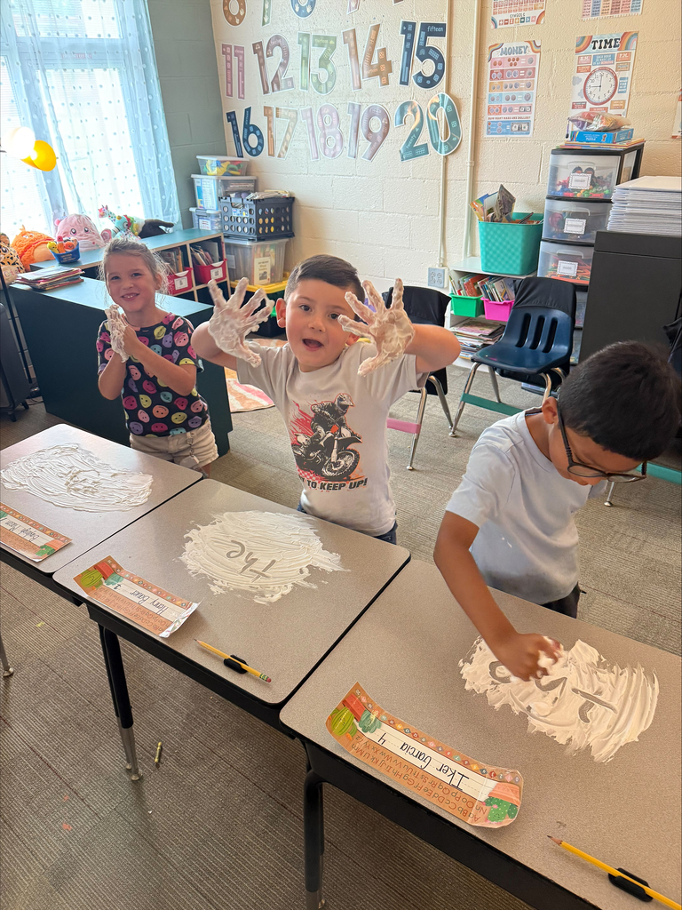 Three students are practicing writing using shaving cream on their desktop.