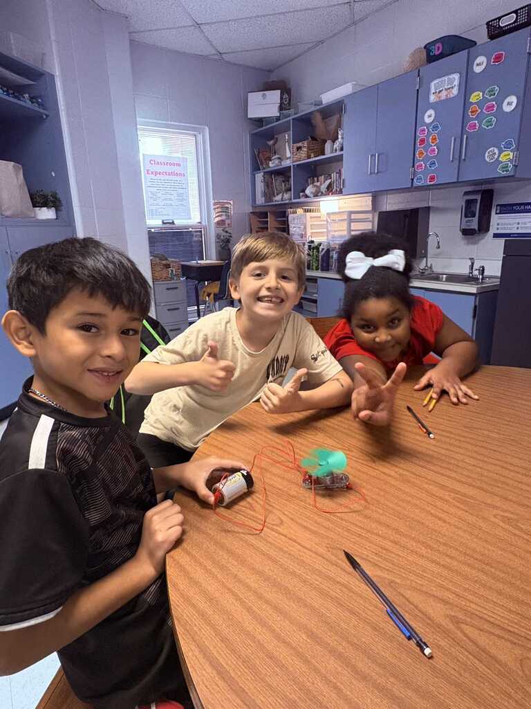 Three children in a classroom, seated at a table. A boy holds a small robot and another has a peace sign.