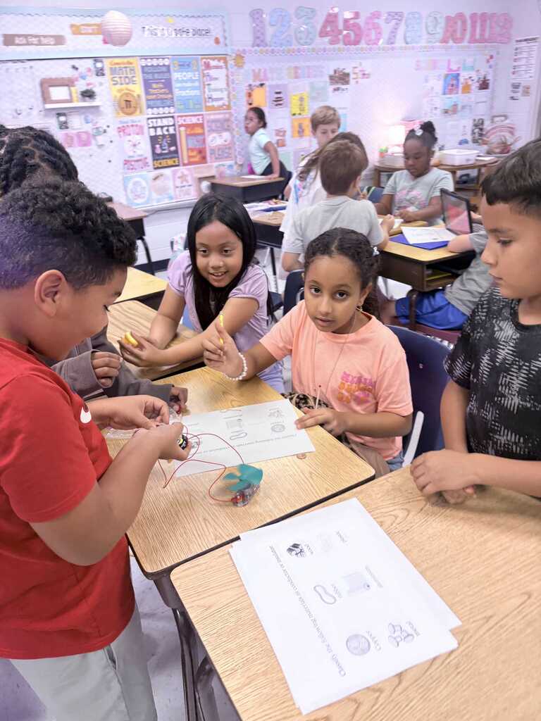 Children work on a project at a table in a classroom with other children and teachers.