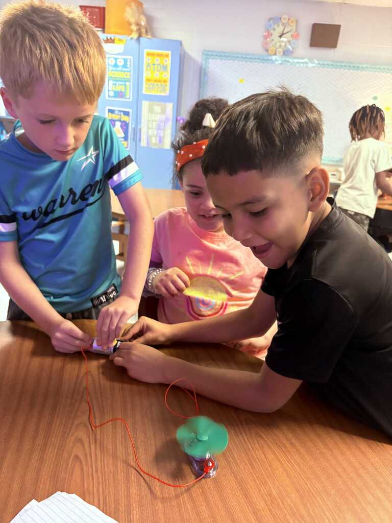 Three kids sit at a table, two boys and a girl, working on a small circuit with wires and a spinning toy.