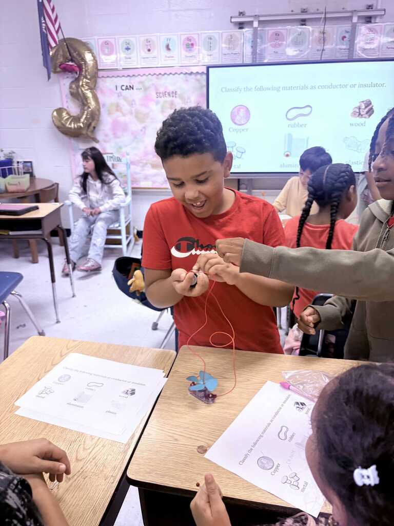 Classroom with students seated at desks; a boy in red holds a wire, another holds his hand.