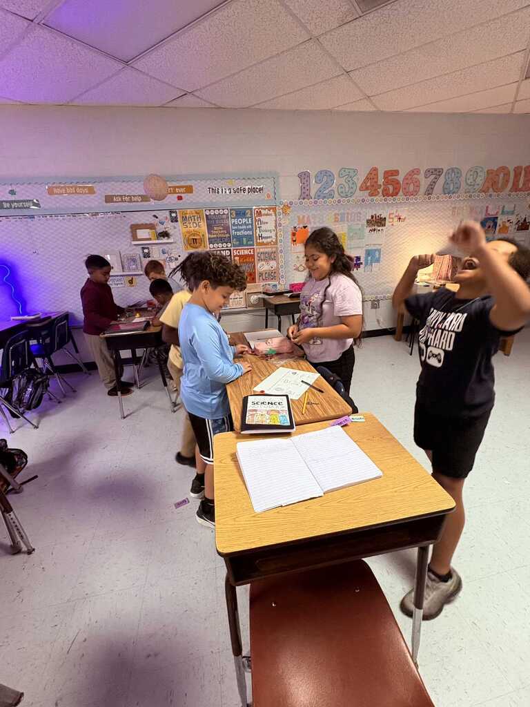 A classroom with children seated at desks, one standing and raising hands. Papers, a tablet, and pens are on a desk.