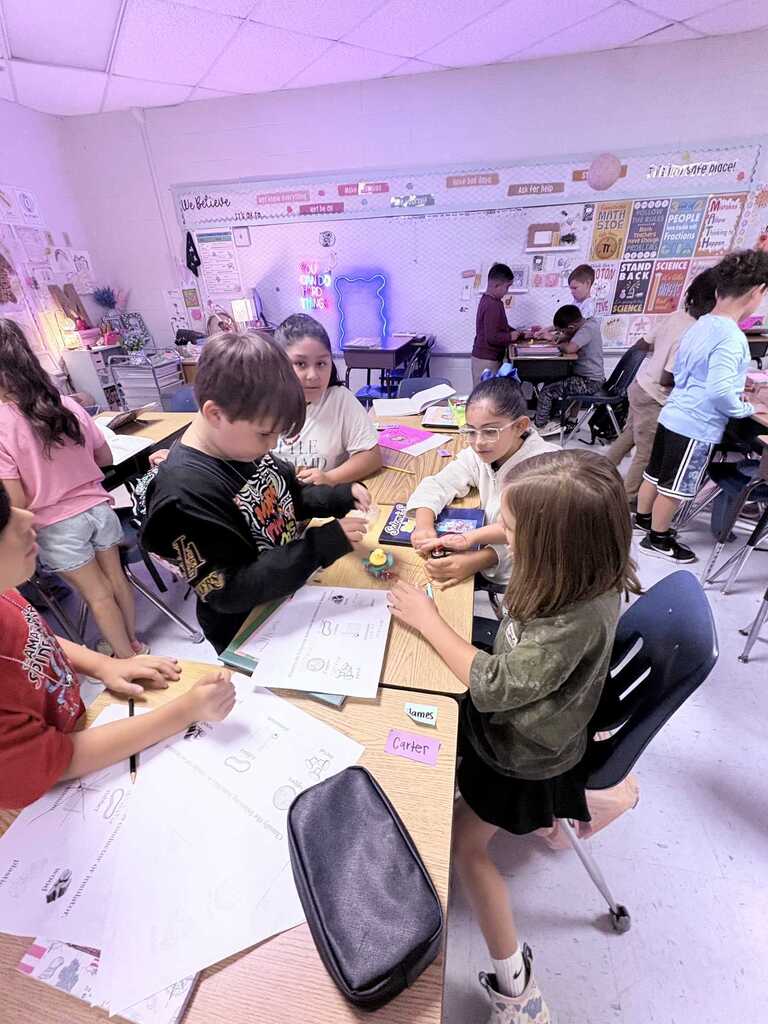 Kids work at a table in a classroom with a whiteboard and other students in the background.