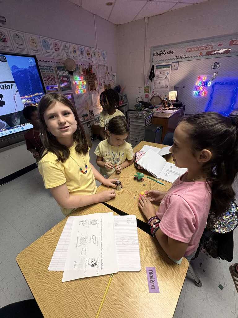 Three young girls seated at a desk in a classroom, looking at a project. One has a necklace, another holds a book.