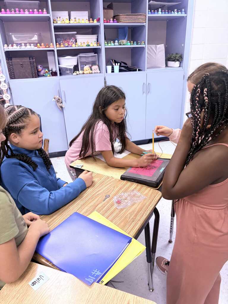 Four girls at a desk, one holding a tablet. Two of them have braids, one in a pink shirt. The background features blue cabinets and shelves.