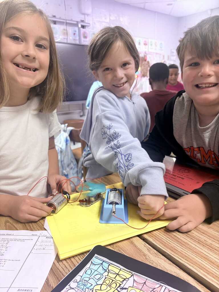 Three children work on a science project together at a desk. They are holding wires and a battery.
