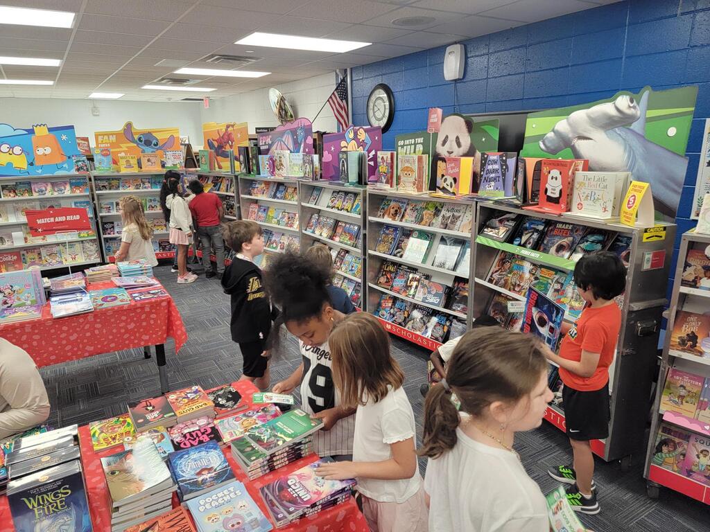 A room with shelves full of books, children looking at them; some books on tables with red covers.