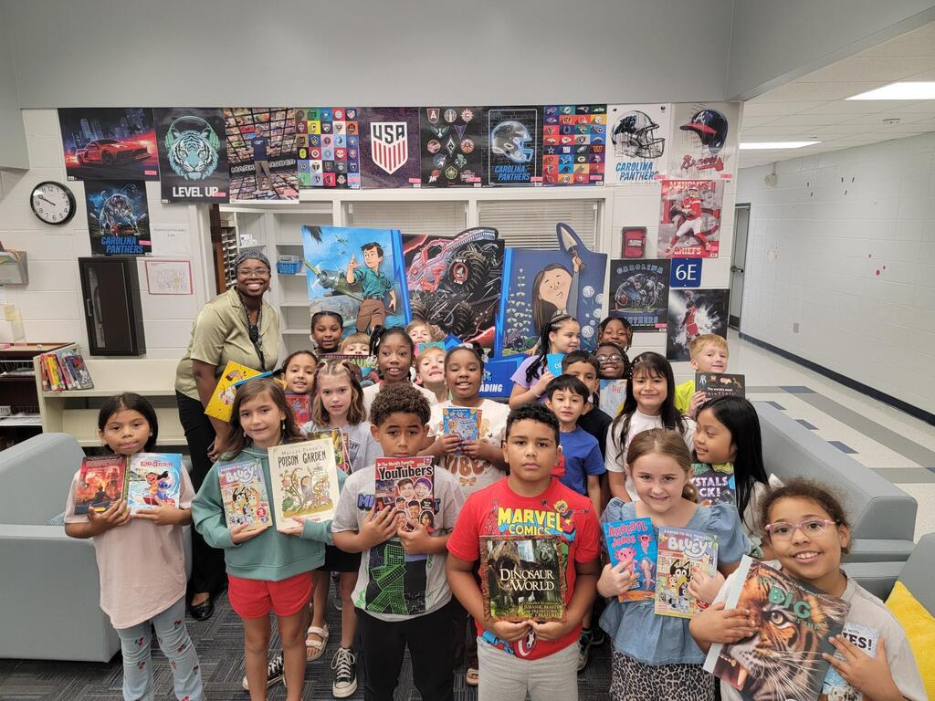 A group of children in a library holding books with a man in the background.