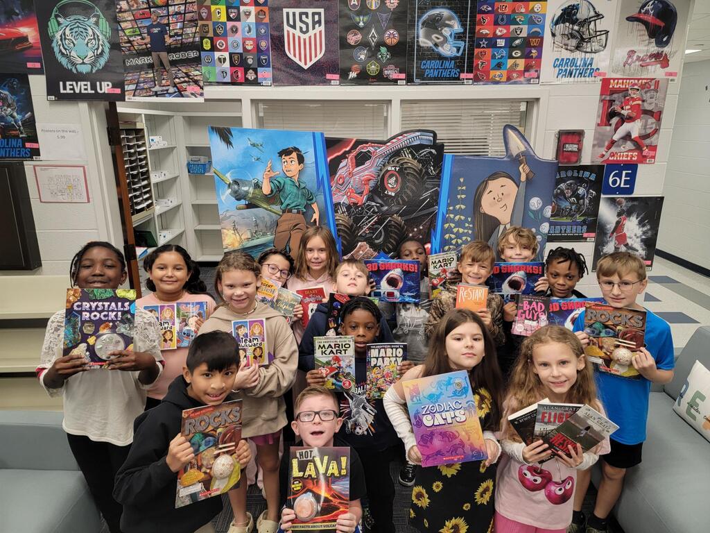 Children in a classroom hold various books, smiling and posing. Background includes posters and a bookshelf with decorations.