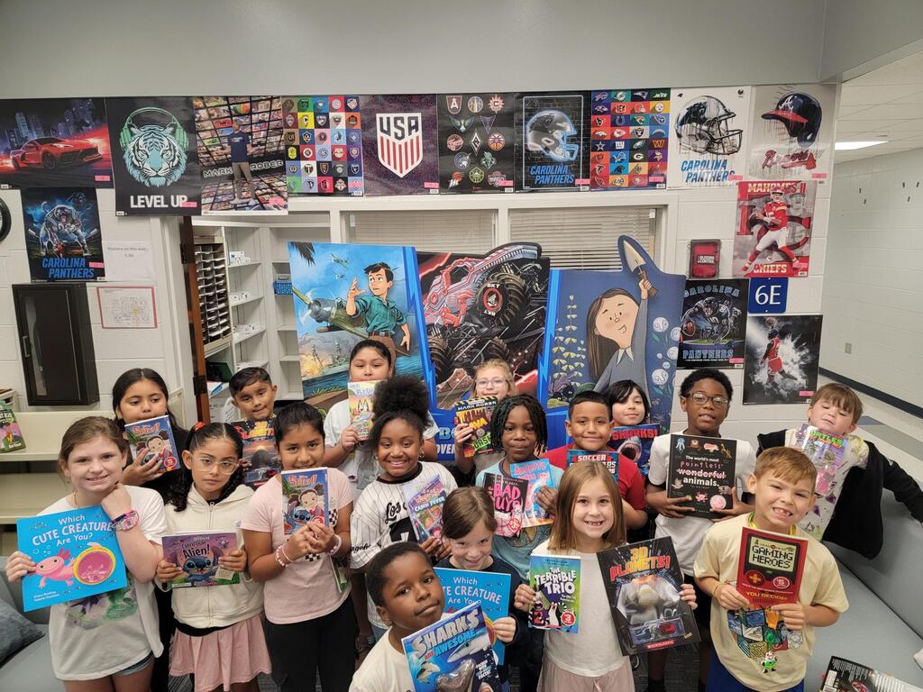 Children in a classroom with bookshelves behind them, holding books and smiling for a picture.