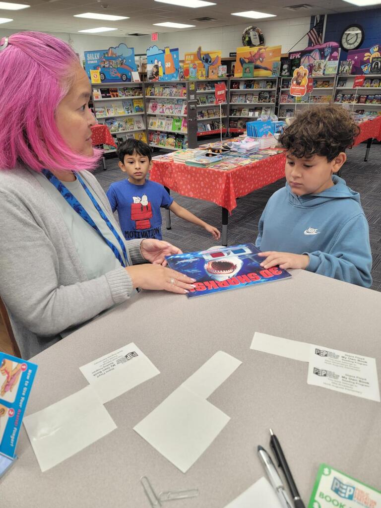 A woman with pink hair sits with two boys at a table, looking at a book and white papers.
