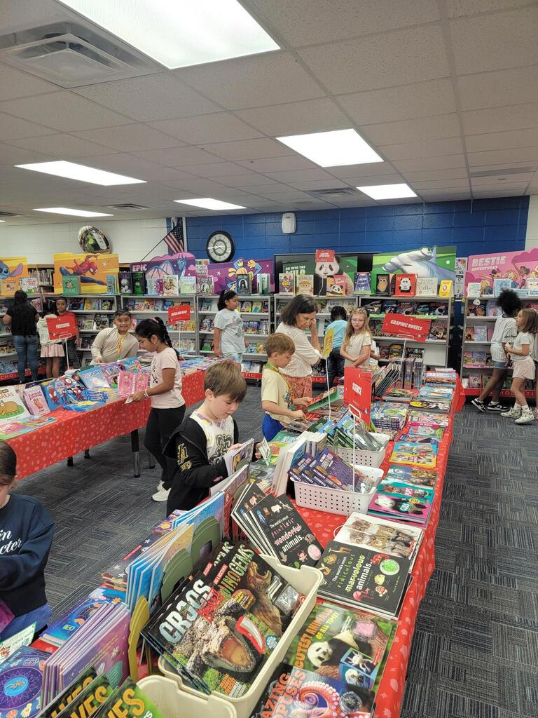 A room with several kids browsing through tables filled with books and toys. Tables have red cloths.