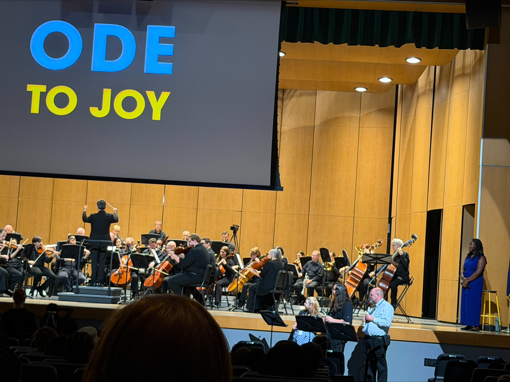 Orchestra playing on stage. A screen above reads "ODE TO JOY". An audience watches from the front.