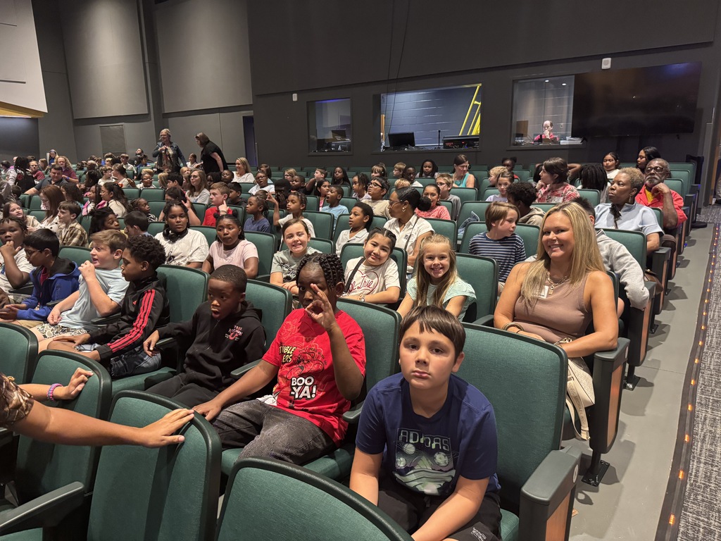 Audience of children and adults in rows of green chairs, seated in an auditorium.