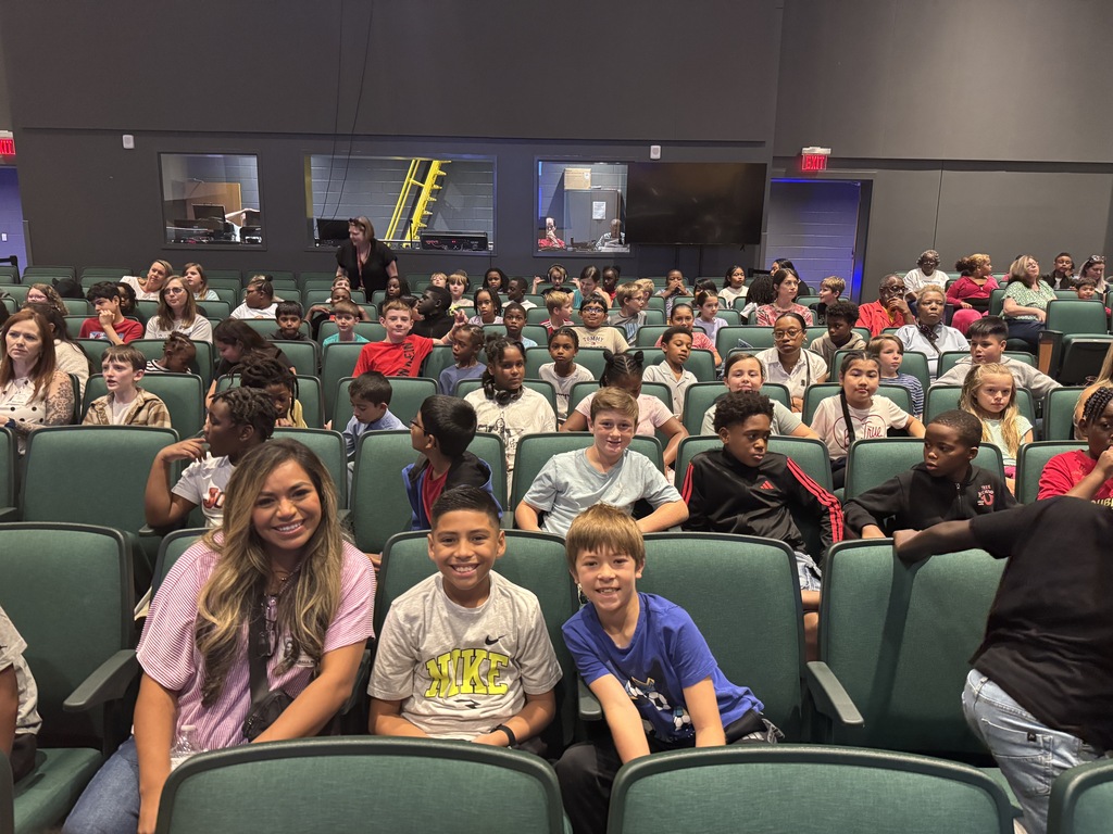 A large group of children and adults in chairs, with one woman smiling at the camera.
