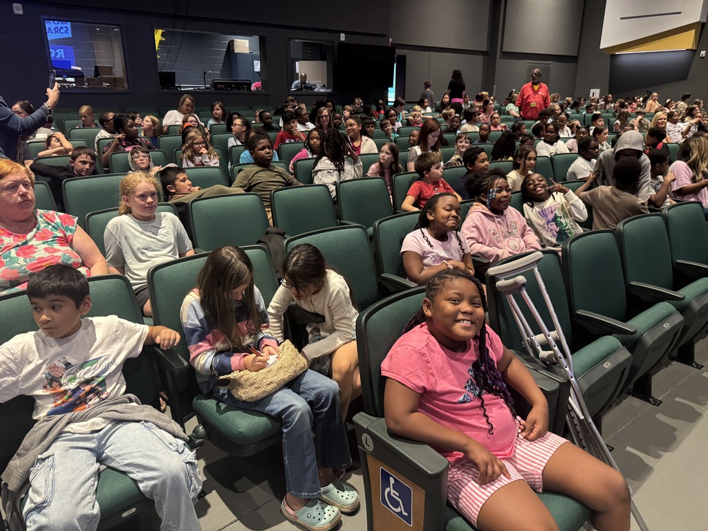 Large crowd in a theater with many rows of green seats; some children are wearing casts.