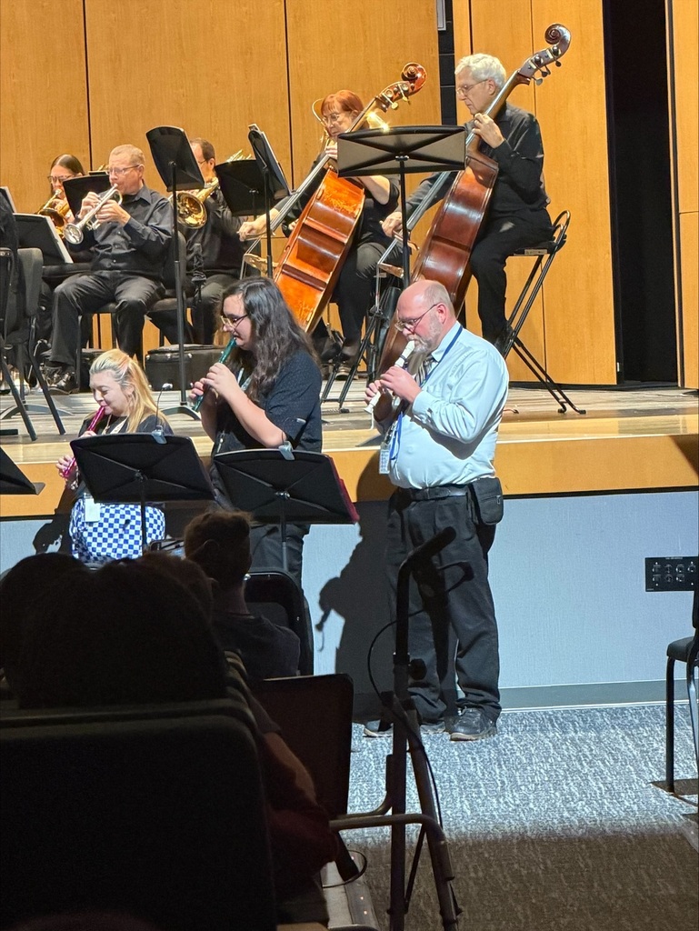 A group of musicians plays instruments on a stage, with audience members seated in chairs in the foreground.