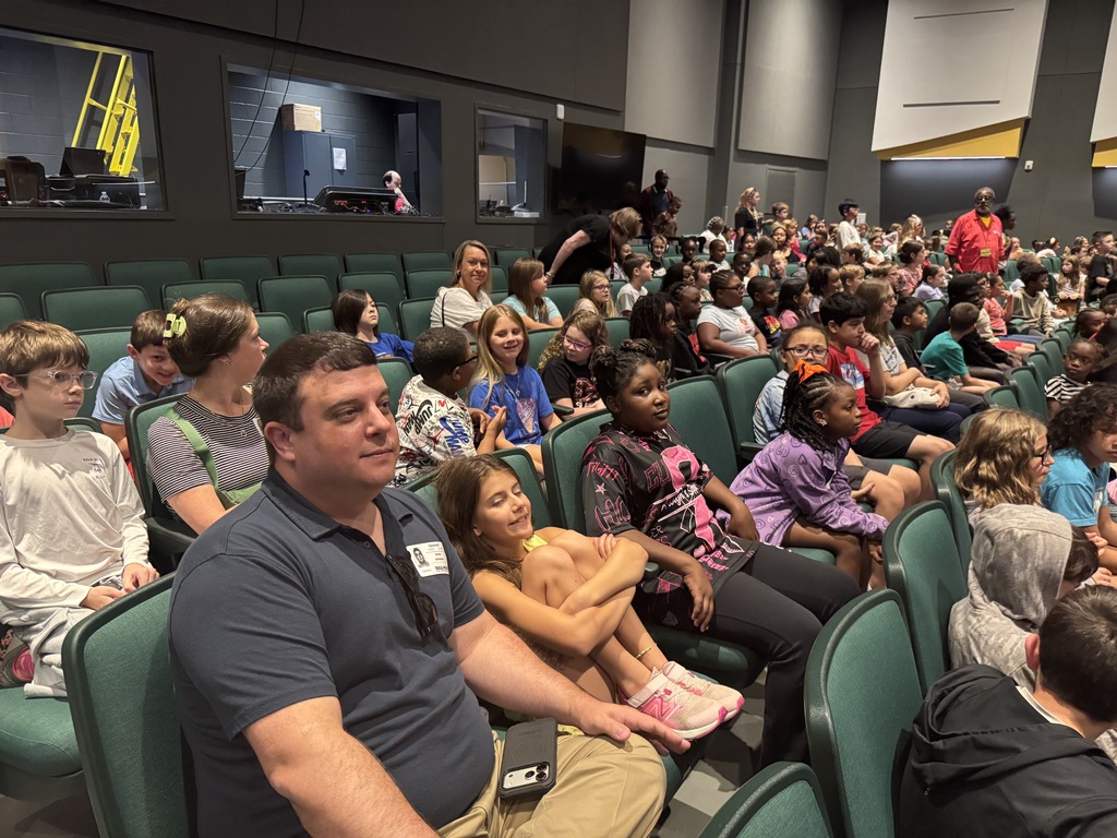 An indoor auditorium filled with children and adults in green chairs. Some children sit on adults' laps.
