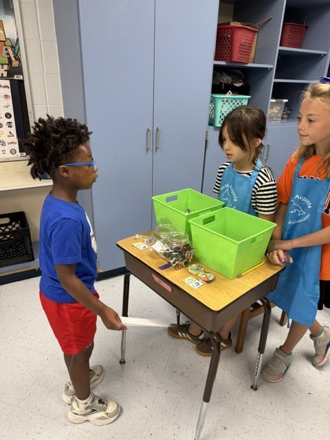 Three children stand at a table with green storage bins on it. A boy in a blue shirt and red shorts holds an object.