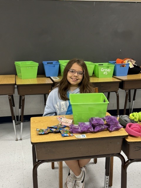A young girl sits at a desk, surrounded by various items like snacks and toys, in a classroom with several desks in the background.