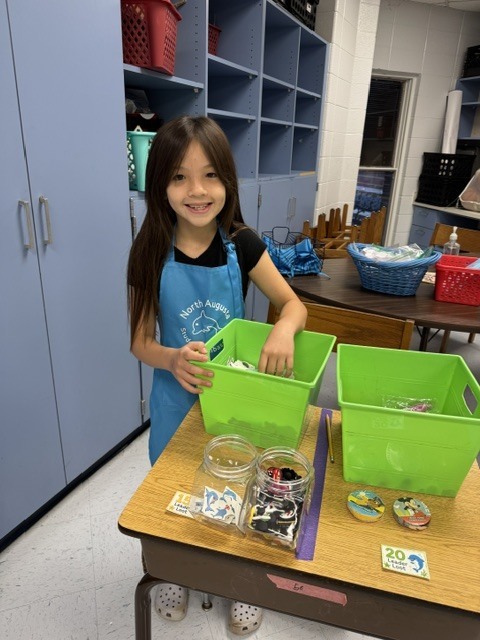 A young girl in a blue apron stands at a table, surrounded by green bins and various items.