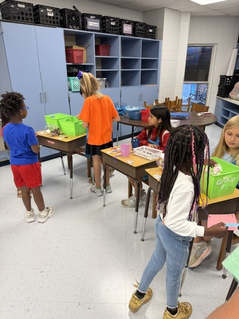 Children stand at desks, engaging in activities. Shelves and cabinets in the background.