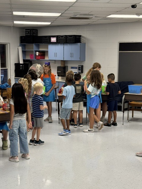 A classroom with children standing in a circle. Overhead lights and cabinets are visible.
