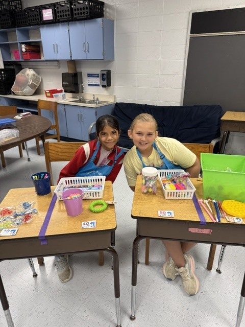 Two children seated at desks, surrounded by art supplies. One has a blue apron, the other a yellow one.