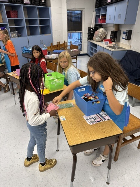 Four girls in a classroom with blue cabinets and desks. One girl has her hand on another's shoulder.