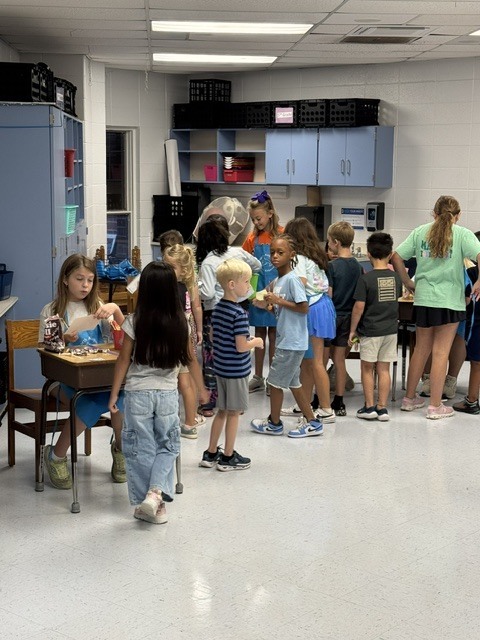 Children and adults gather in a room with cabinets and desks. Several children are seated, and others stand near the seated ones.