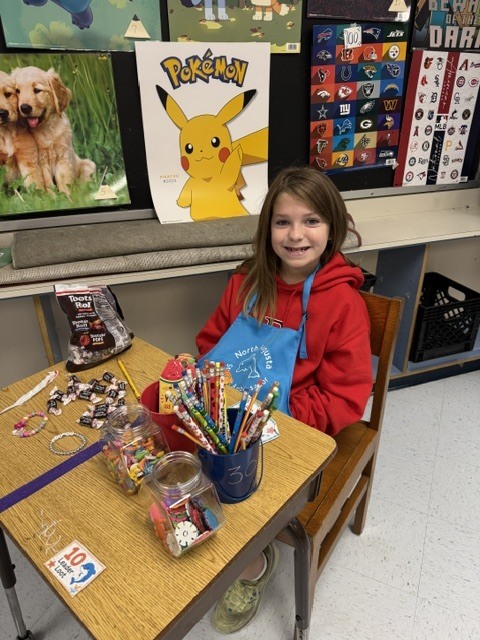 Girl in a red hoodie sitting at a table surrounded by colorful art supplies and posters.