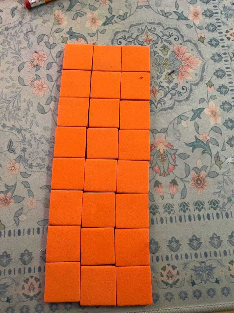 A stack of orange square blocks on a floral-patterned surface with a small, yellow marker nearby.