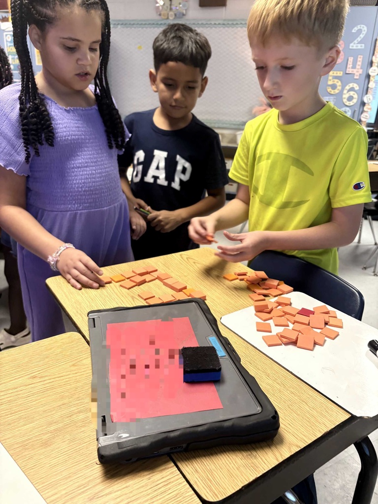 Three children are in a classroom. They are seated at a table, arranging orange tiles. A digital tablet is also on the table.