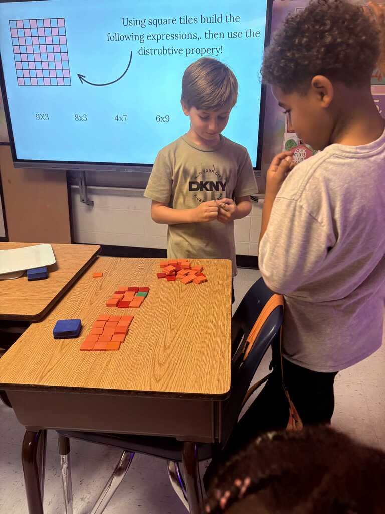 Two boys in a classroom setting. One arranges tiles on a desk, and the other watches. Behind them, a large screen displays a mathematical problem.