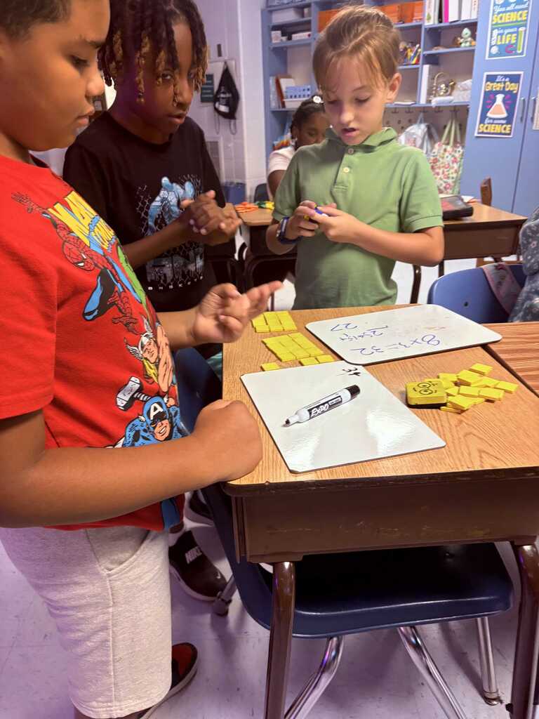 Four children interact at a desk, focusing on a board with colorful tiles and a marker. They are in a classroom setting with shelves and chairs.